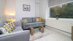 Un salon moderne avec un canapé gris, une table basse en verre et une vue sur les arbres par une grande fenêtre.