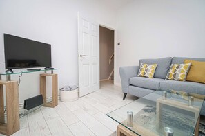 A living room with a flat-screen TV, glass coffee table, grey sofa with patterned cushions, and a wooden door leading to another room.