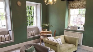 A room with a green wall, a green sofa, a brown armchair, a small table, and a window with a view of greenery. - The Cornish Peak: Cornwall by STAE-Homes (Cornwall)