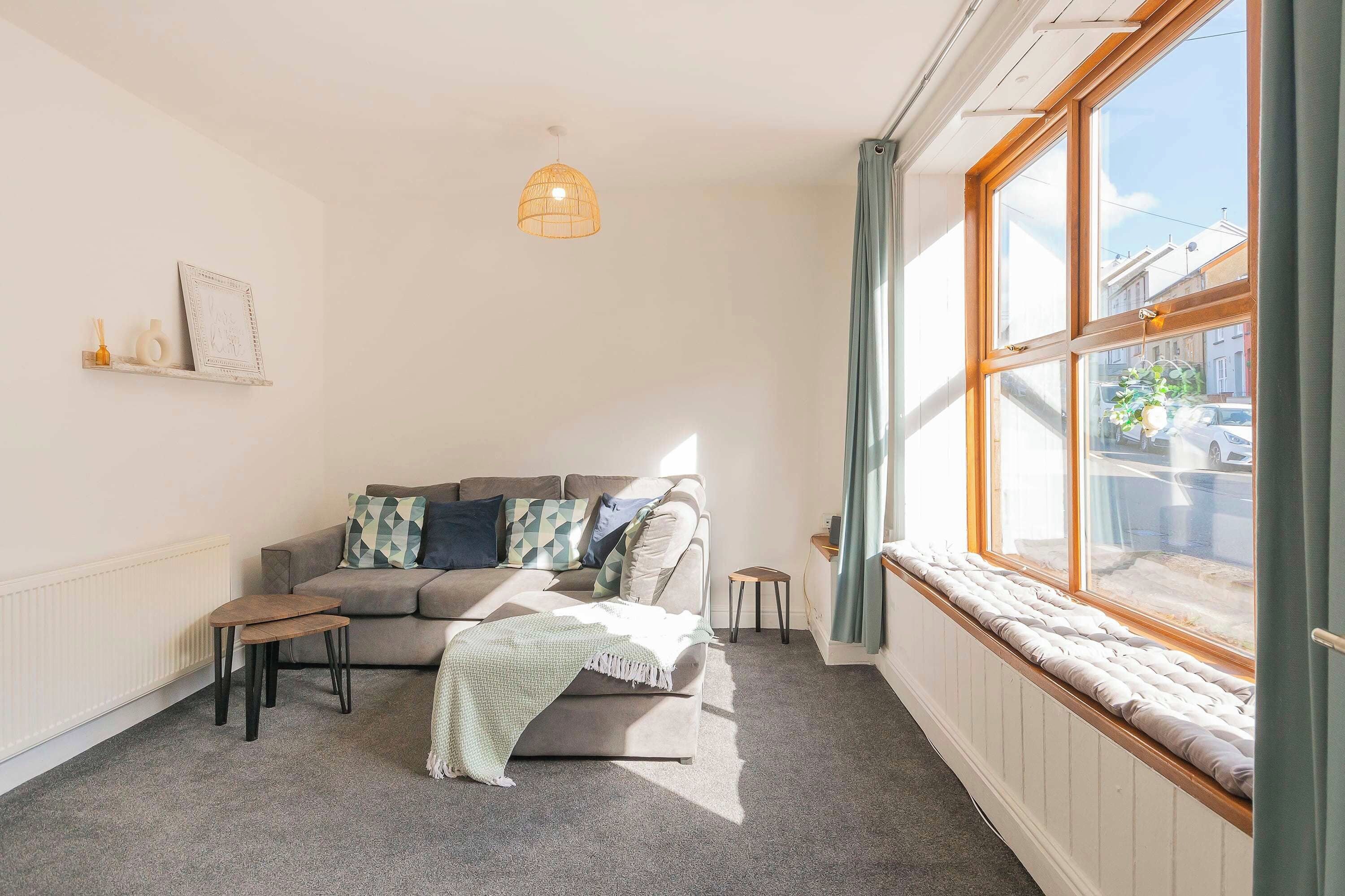 A cozy living room with a sofa, a small table, a hanging light fixture, and a window with curtains.