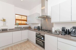 Una cuina moderna amb armaris blancs, una campana extractora d'acer inoxidable, un forn integrat i una cafetera.