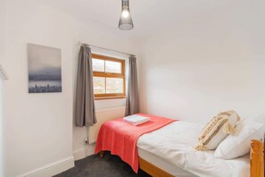 Une chambre avec un lit simple, un radiateur, une fenêtre avec des rideaux, un tableau accroché au mur et un luminaire suspendu.