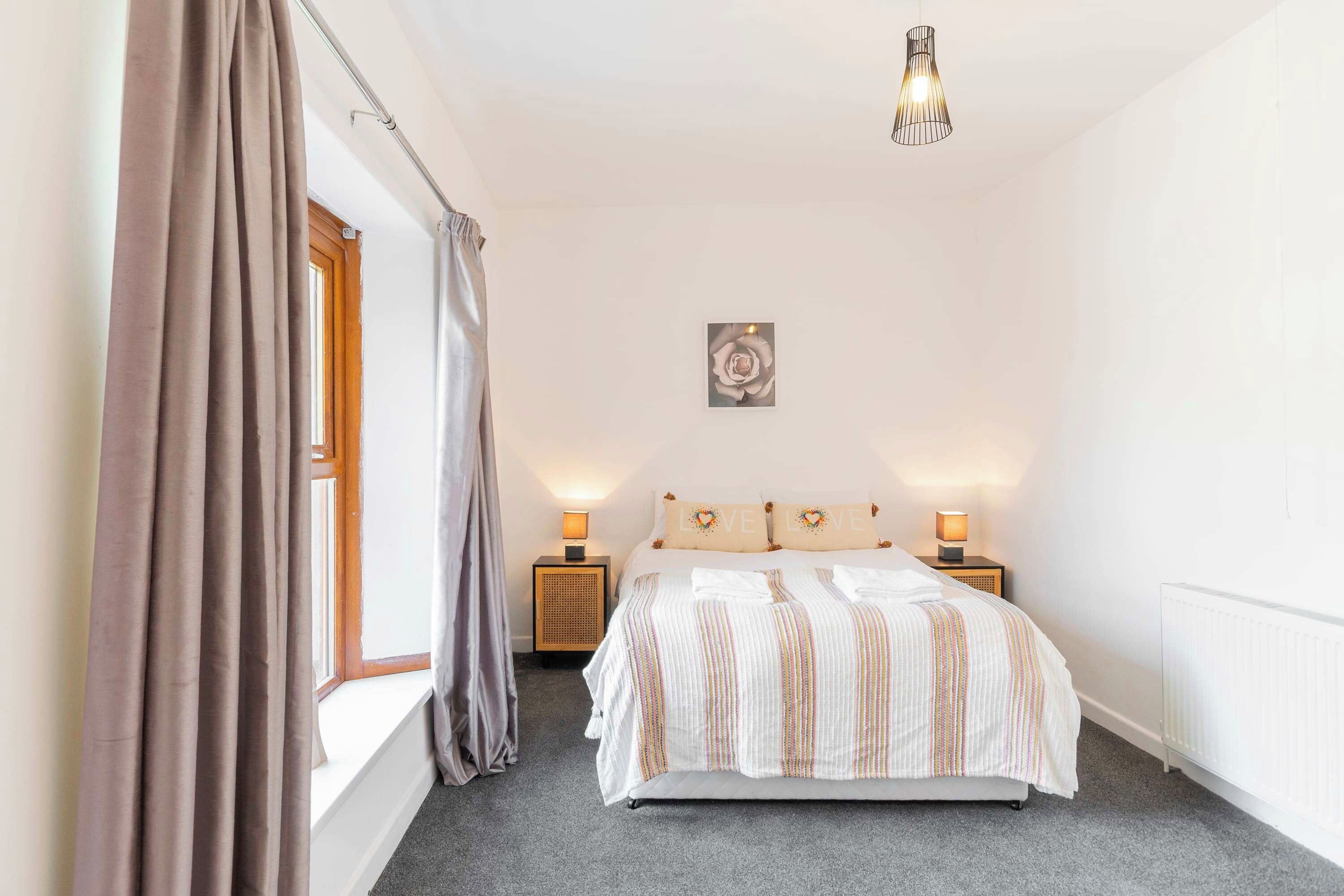 A bedroom with a bed, bedside lamps, a window with curtains, a wall-mounted picture, and a ceiling light fixture.