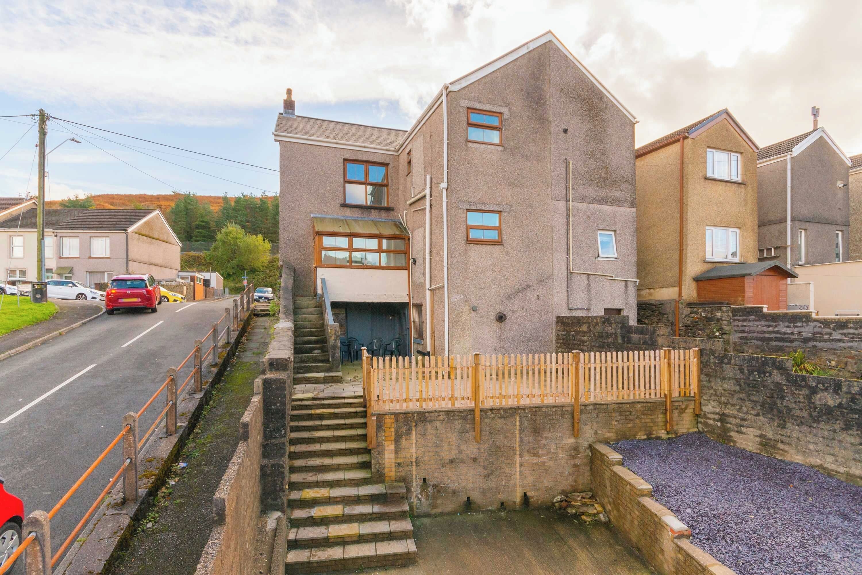 A residential building with a staircase leading to the entrance, a gated driveway, and a road with parked cars.