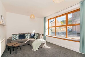 A cozy living room with a grey sofa, a small wooden table, and a large window with curtains.
