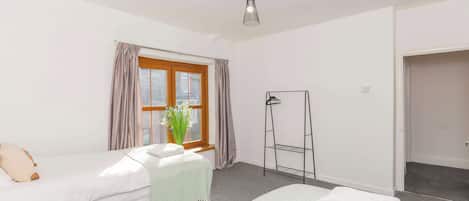 A hotel room with two beds, a window with curtains, a plant, a black metal shelf, and a door leading to another room.