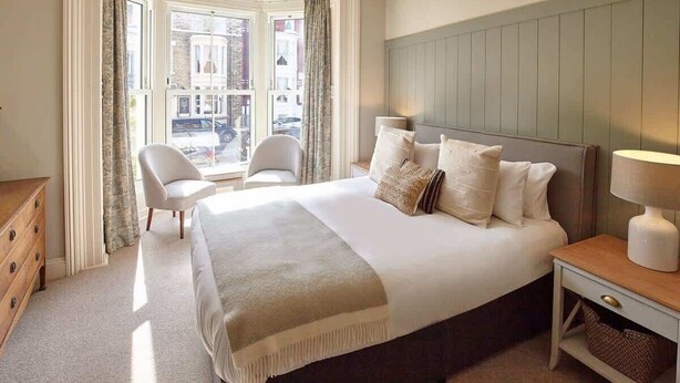 Quarto de hotel com uma cama grande, uma cadeira, uma mesa de cabeceira e vista para a rua pela janela.