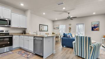 A modern kitchen with white cabinets, a built-in microwave, and a dishwasher. There is a dining area with a blue sofa and a striped armchair.