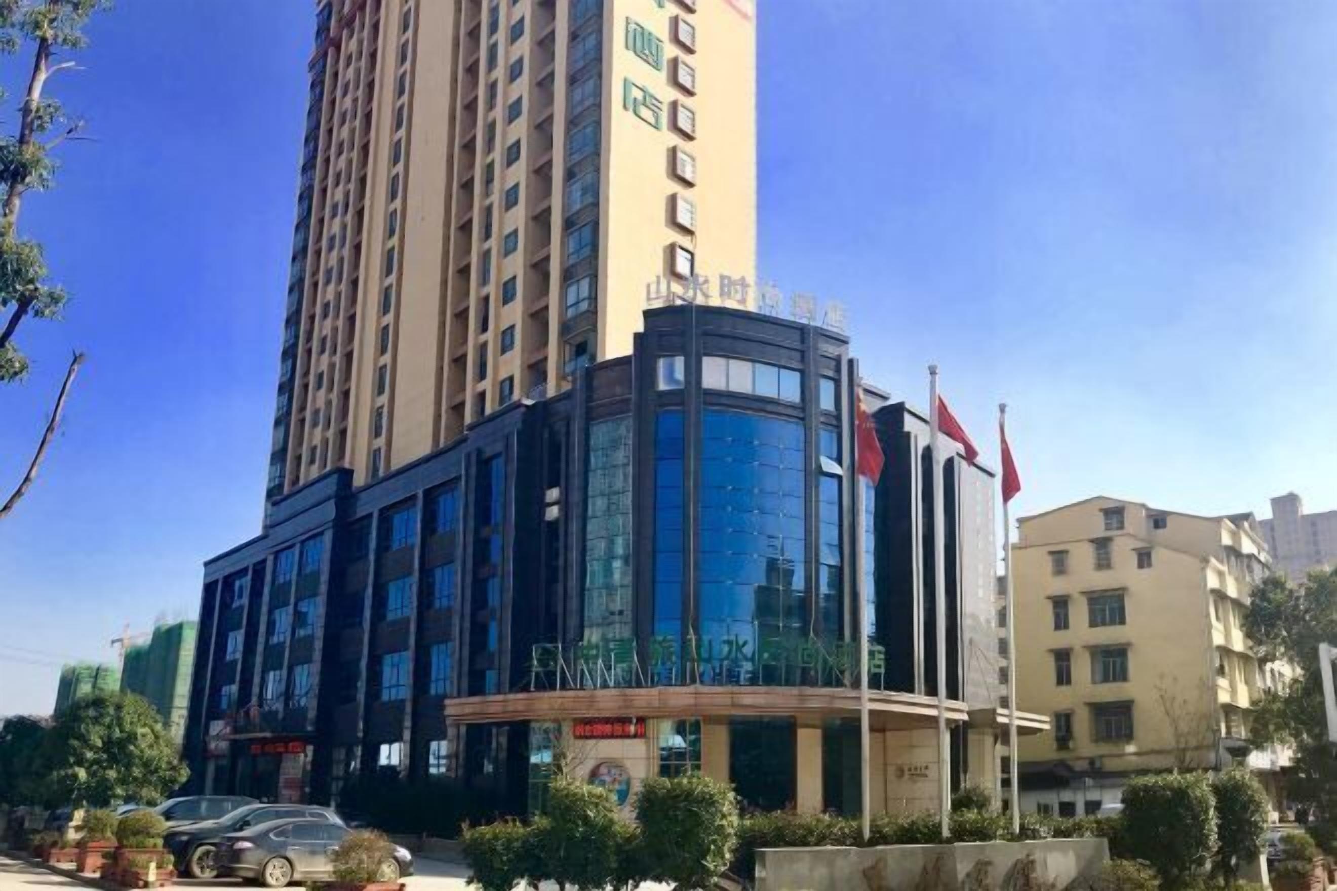 A modern hotel with a curved glass facade, a flagpole, and a parking lot with cars.