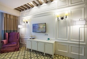 A hotel room with a television, a cushioned armchair, a white console table, and patterned floor tiles.