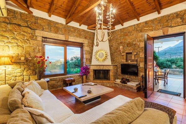 A cozy living room with a stone fireplace, a television, and a large window offering a view of the outdoors.