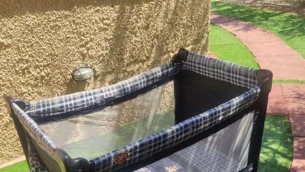 A portable crib with a checkered fabric and mesh sides, placed on artificial grass near a stone wall.