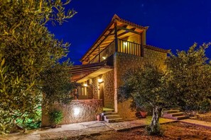 Een stenen villa met een houten balkon, een stenen inkom en een goed verlicht buitengebied 's nachts.