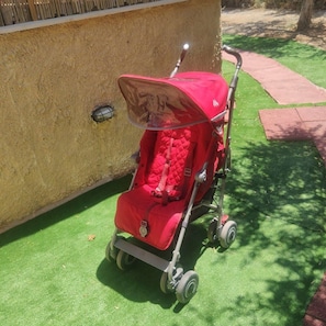 Ein roter Kinderwagen mit Verdeck auf künstlichem Rasen neben einer Mauer.