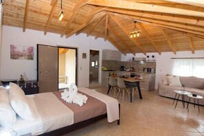 Een ruime kamer met een hoge nok, een bed met witte lakens, een eethoek met een tafel en stoelen, en een kitchenette op de achtergrond.