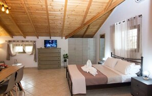 Een slaapkamer met een houten plafond, een bed met witte lakens, een televisie op een dressoir en een bureau met stoelen.