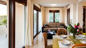 A dining area with a table set for four, a sofa in the background, and large windows with curtains. - Armonia by the Sea - Majestic Seaside Retreat (Petalidi)