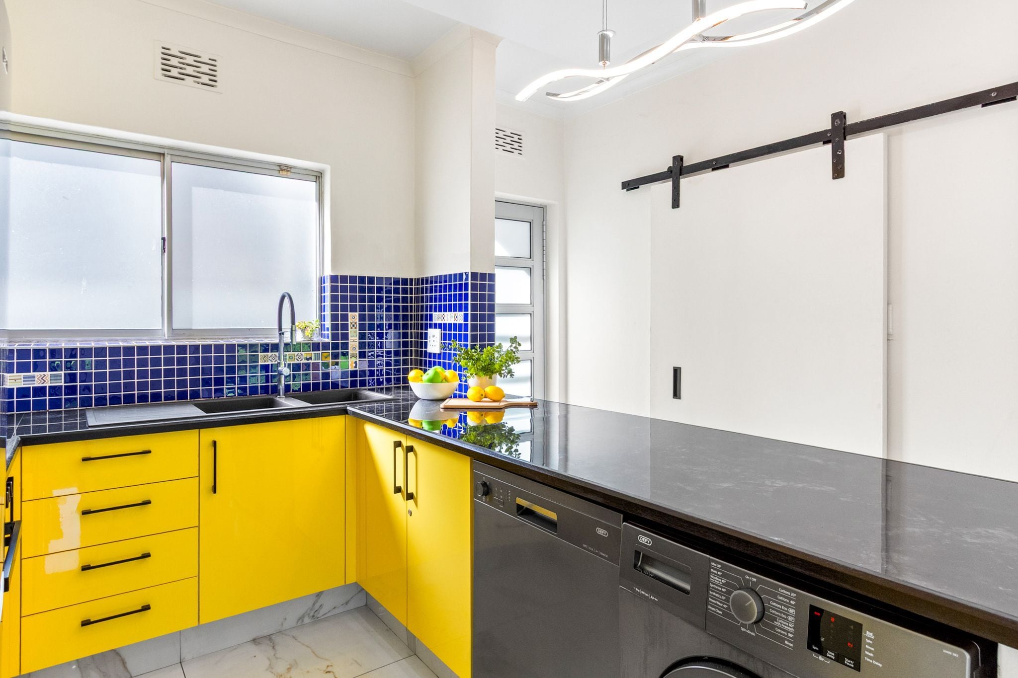 A kitchen with yellow cabinets, a black countertop, and a window with blue-tiled backsplash.