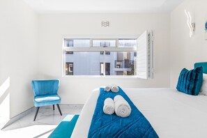 A hotel room with a large bed, a blue chair, and a view of buildings through a window. - Manella On Kloof (Cape Town)