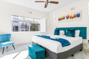 Une chambre moderne avec un grand lit, une chaise bleue, un banc turquoise et un ventilateur de plafond.
