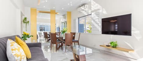 A modern living room with a sofa, dining table, and chairs. There is a TV mounted on the wall and a plant on a shelf.