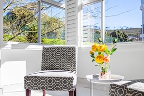 A modern living room with a patterned armchair, a small round table with a vase of yellow flowers, and a view of greenery and buildings through large windows.