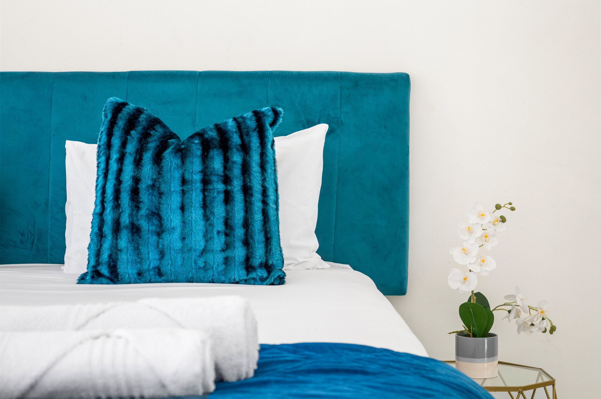 A bed with a teal headboard, white pillows, a blue blanket, and a white orchid in a pot.