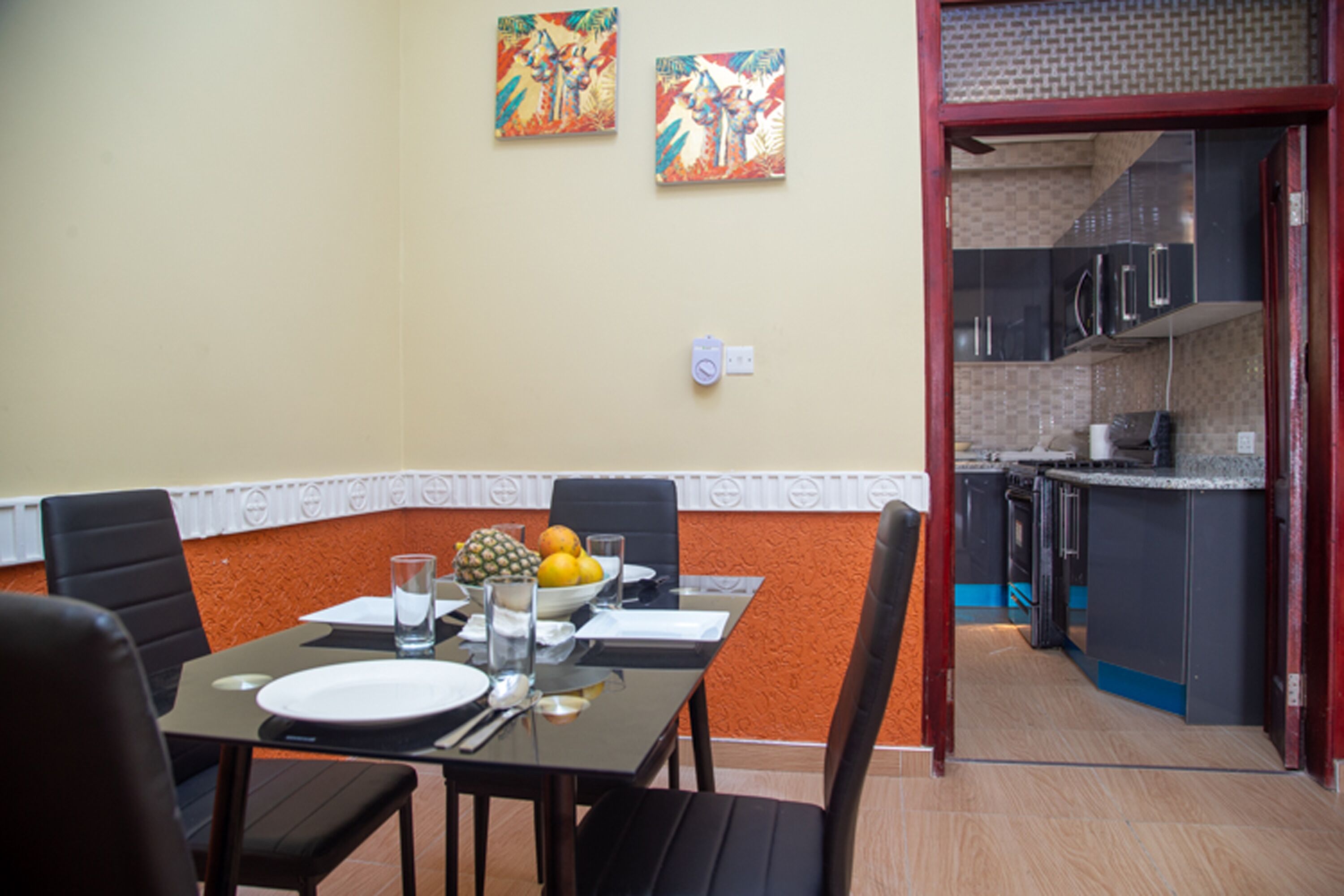 A dining area with a table set for four, a fruit bowl, and two wall-mounted paintings.