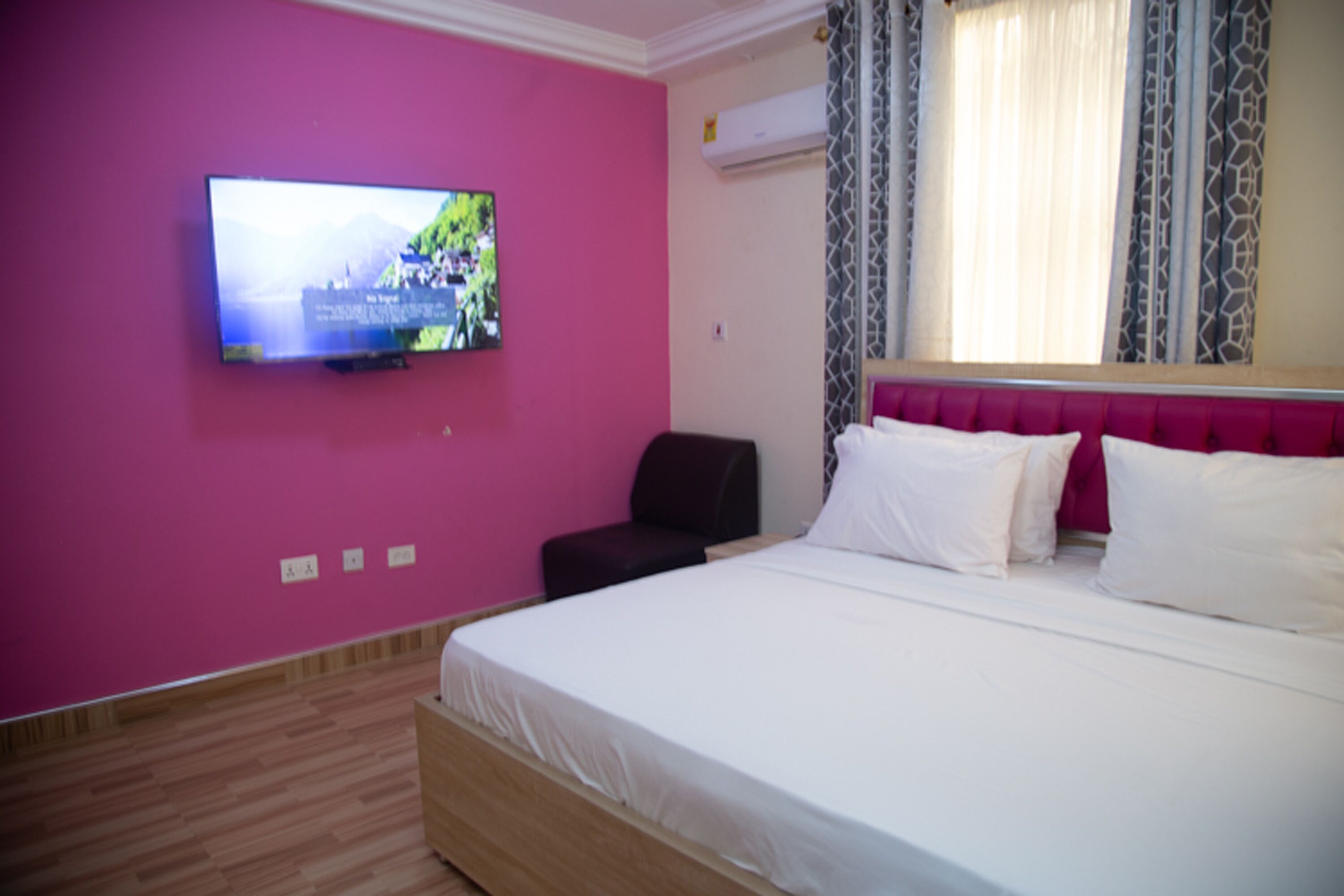 Ein Hotelzimmer mit einem Bett, einem an der Wand befestigten Fernseher, einem Sessel und einem Fenster mit gemusterten Vorhängen.