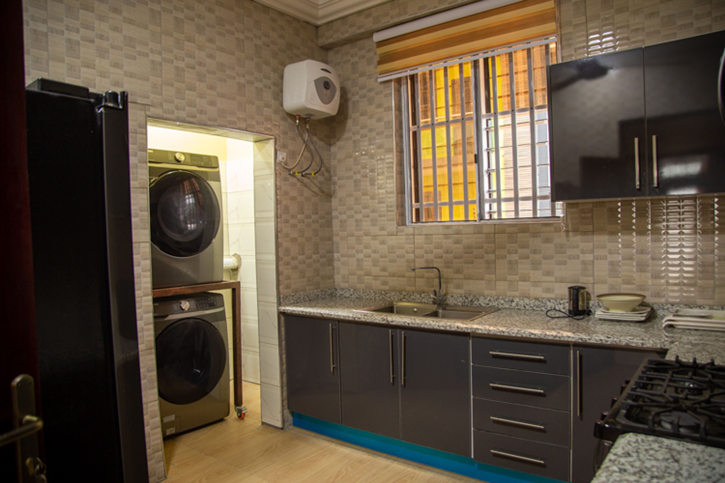 Une cuisine compacte équipée d’un lave-linge et d’un sèche-linge intégrés, d’une cuisinière à gaz et d’une fenêtre avec des stores.