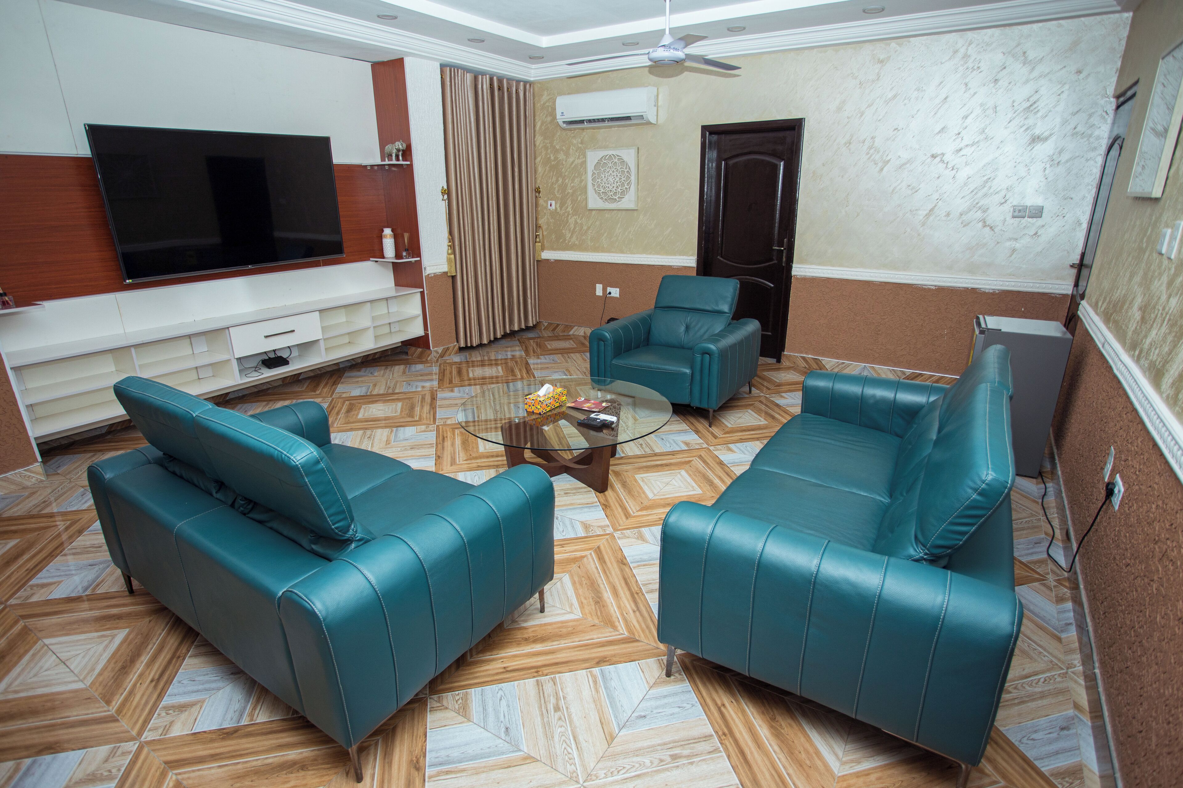 A living room with a flat-screen TV, two teal leather sofas, a coffee table, and patterned flooring.