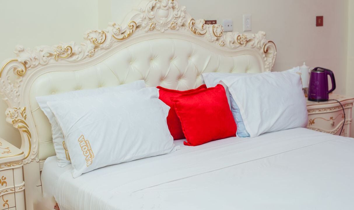 A bed with white and red pillows, a white headboard with gold detailing, and a purple kettle on a bedside table.