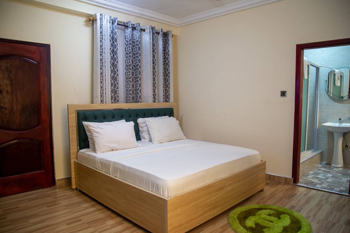 Un dormitorio con cama de madera, ventana con cortinas estampadas, puerta de madera y baño con lavamanos y espejo.