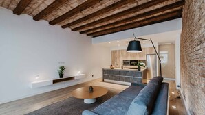 A modern living room with a grey sofa, a round coffee table, and a pendant light. A kitchen is visible in the background with wooden cabinets and a stainless steel refrigerator. - Petit Luxe Sant Pere (Terrassa)