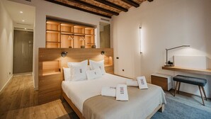 A modern hotel room with a wooden headboard, a large bed with white linens, a desk with a lamp, and a bench. - Petit Luxe Sant Pere (Terrassa)