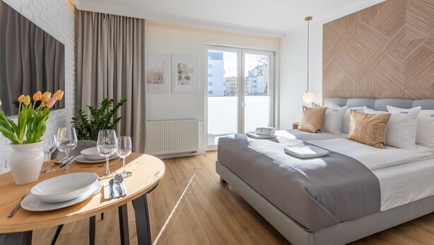 A modern hotel room with a large bed, a dining table with plates and glasses, and a view of the city through the window.