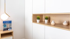 A minimalist room with a wooden shelf holding potted plants and vases, a desk with a lamp, and a book titled "Ambra".