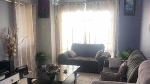 A living room with a chandelier, a sofa, a coffee table, and a television. - Stay.Plus Kahawa Sukari Apartment (Ruiru)