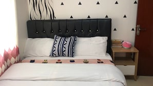 A bed with a black upholstered headboard, white bedding, and a patterned pillow. There is a wall with black triangle decals and a decorative wall hanging. A small wooden nightstand with a pink object and a door are also visible. - Stay.Plus Kahawa Sukari Apartment (Ruiru)