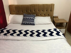 A bed with a tufted headboard, two pillows, a patterned blanket, and a bedside table with books. - Stay.Plus Kahawa Sukari Apartment (Ruiru)