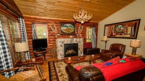 A cozy log cabin interior with a stone fireplace, leather furniture, and a mounted television. - The Log Cabin at PeaceRidge (Wilmington)
