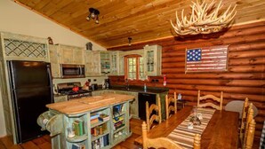A rustic cabin interior with a wooden dining table, a kitchen area with a refrigerator, microwave, and cabinets, and a mounted American flag. - The Log Cabin at PeaceRidge (Wilmington)