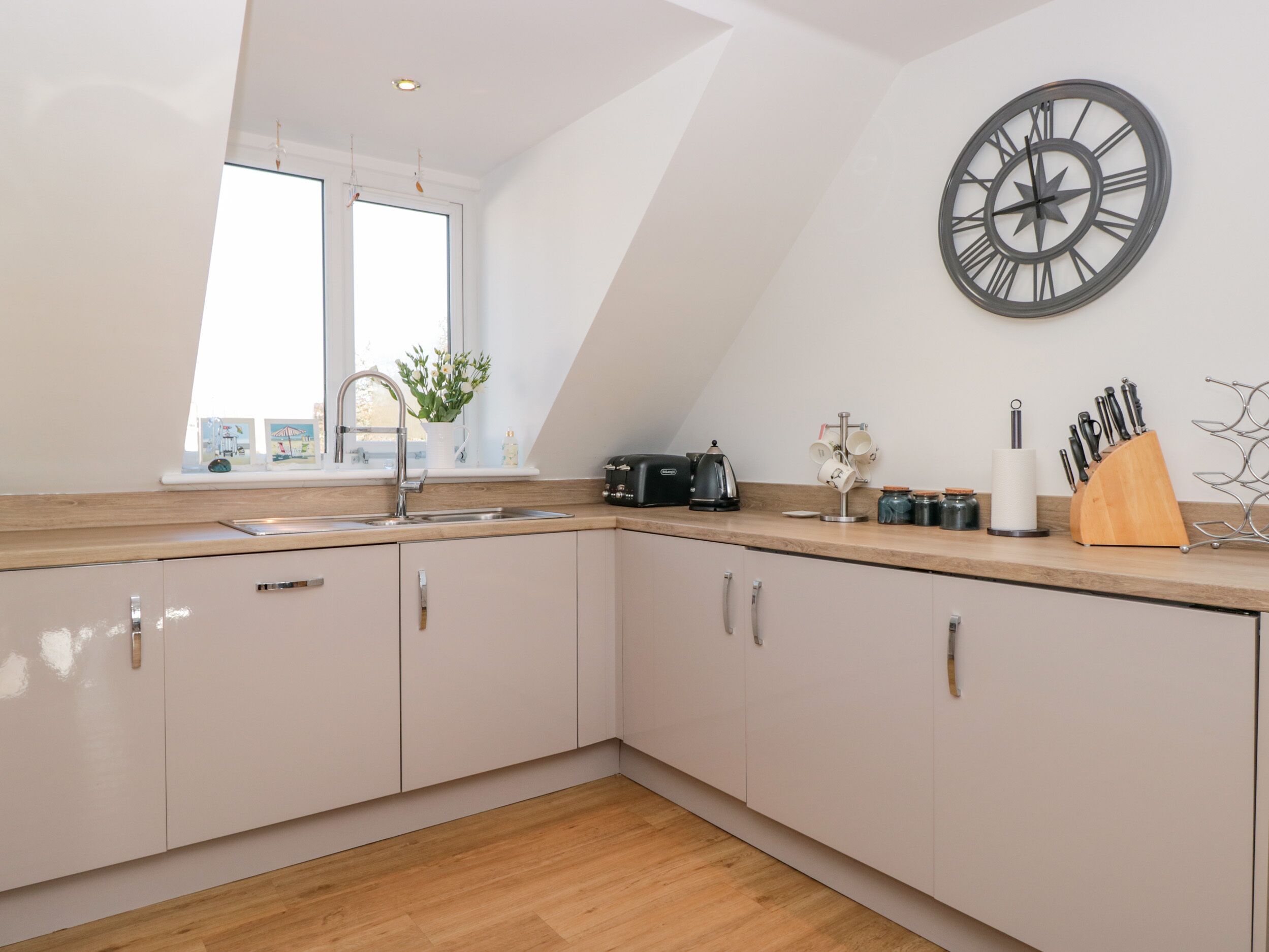 A modern kitchen with a wooden countertop, white cabinets, a large wall clock, and a window with a view.