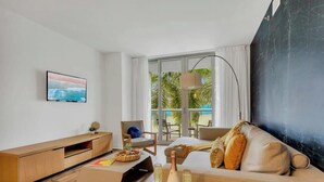 A modern hotel room with a large window, a flat-screen TV, a sofa, a chair, and a lamp. - Apartment w/ Balcony+ Pool+ Near Beach (Hallandale Beach)