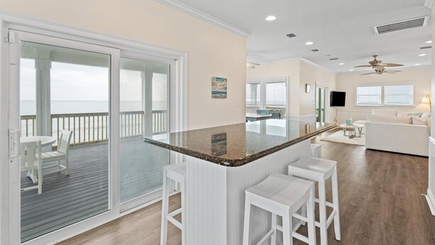 A modern kitchen with a large island, sliding glass doors to a balcony, and a seating area with a white sofa and chairs. - Shamrock Shores - Gulf Front West End Pet Friendly Property With Room For Everyone. 3 Master Suites! 6 Bedroom Home by RedAwning (Dauphin Island)