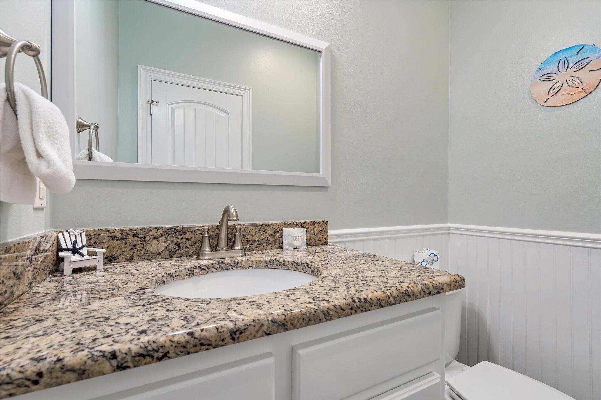 A bathroom with a granite countertop sink, a white vanity, a towel rack, and a decorative wall piece.