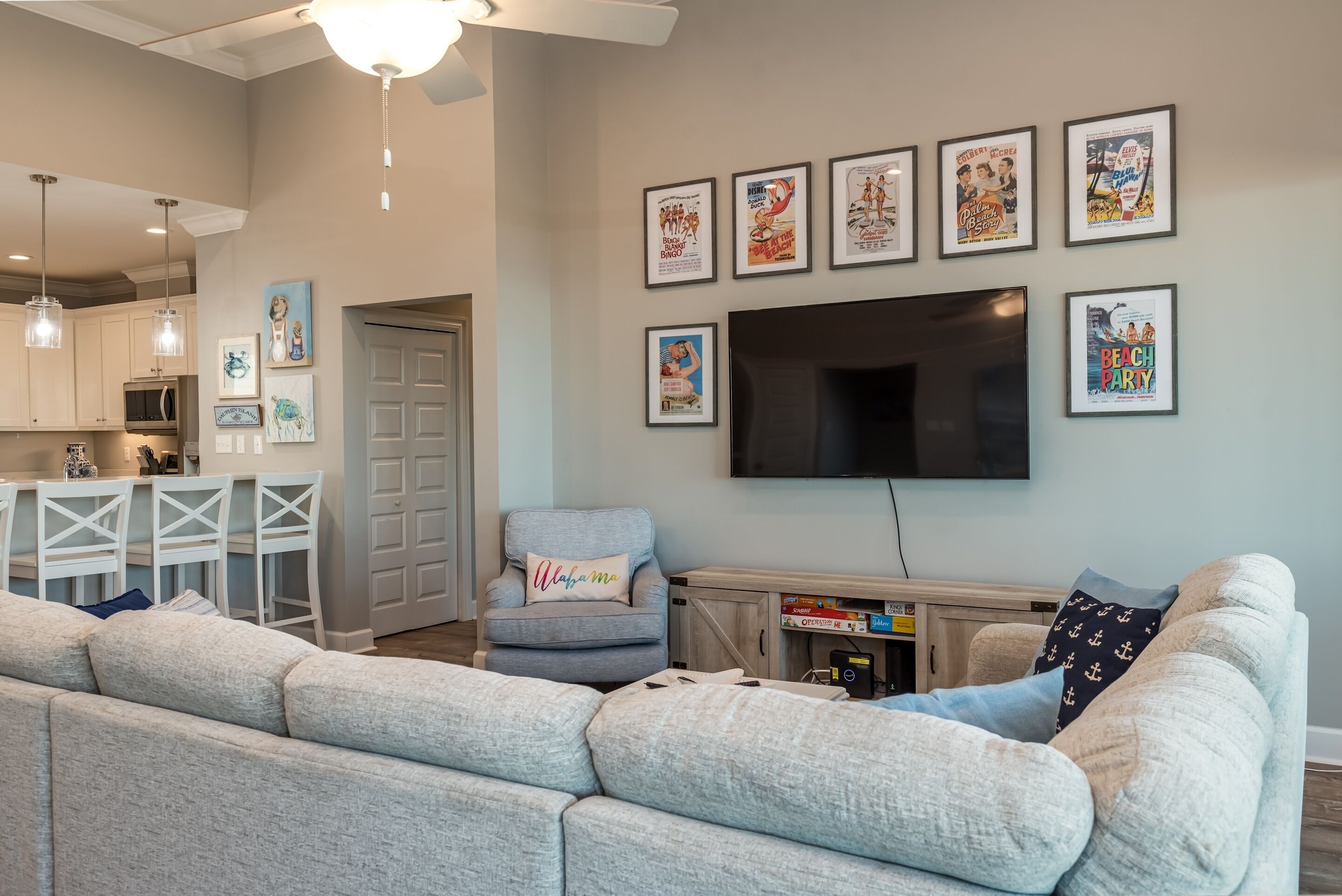 A living room with a gray sectional sofa, a gray armchair, a flat-screen TV mounted on the wall, and a kitchen area with white cabinets and pendant lights.