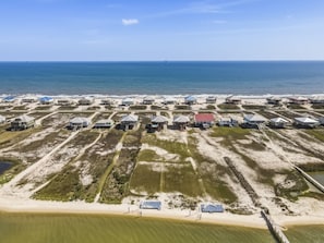 An aerial view of a coastal area with multiple beachfront properties, including houses and buildings with roofs, some elevated on stilts, and a sandy beach in the foreground. - Blue Oasis - Private Backyard Beach - Pool, Hammocks, Kayaks, Even Crab Traps So You Can Catch Dinner! 5 Bedroom Home by RedAwning (Dauphin Island)