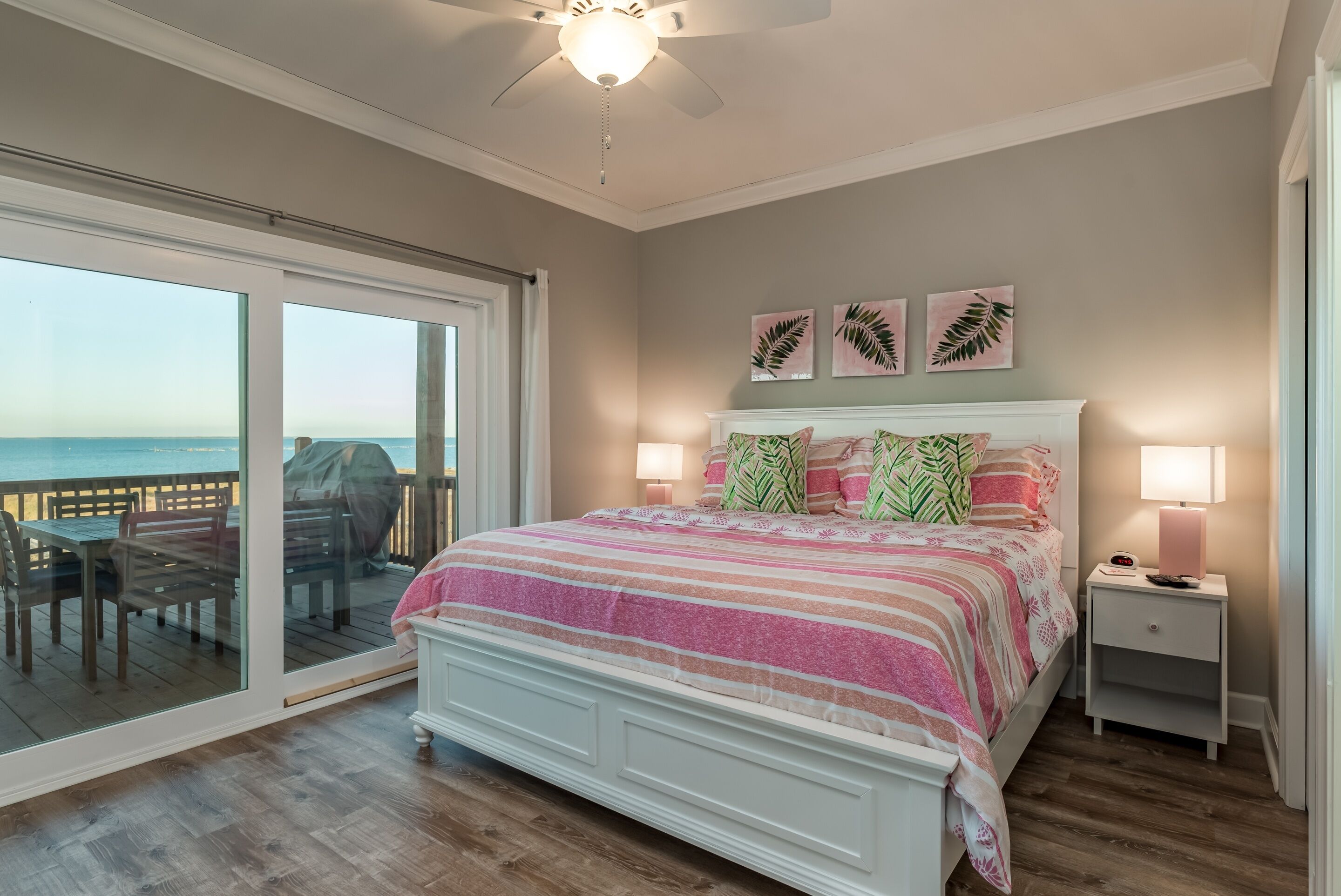 A bedroom with a large sliding glass door leading to a balcony, a bed with a striped blanket, two wall-mounted lamps, and three leaf-patterned wall art pieces.