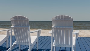 Zwei weiße Adirondack-Stühle auf einer Holzterrasse am Meer.